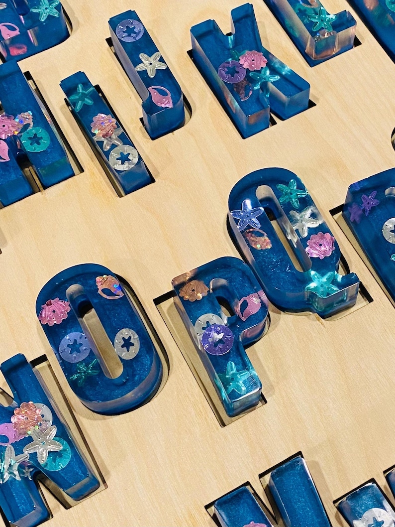 A close-up of a wooden puzzle board, showing several chunky, translucent blue resin letters with embedded glitter and seashell shapes, fitting into the board's cutouts.