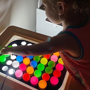 A child's hand reaches into a wooden table with a black acrylic insert holding 32 colorful, translucent ping pong balls, lit from below.