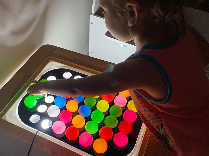 A child's hand reaches into a wooden table with a black acrylic insert holding 32 colorful, translucent ping pong balls, lit from below.