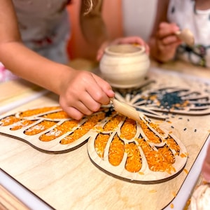 Op de afbeelding: Een houten vlinder-vormig dienblad met oranje en zwarte rijst die de uitgesneden ontwerp vult. Een kinderhand gebruikt een houten gereedschap om het dienblad met rijst te vullen.