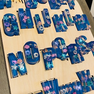 A wooden puzzle board with 26 chunky, translucent blue resin letters, each embedded with glitter and small seashell shapes, filling the letter-shaped cutouts.