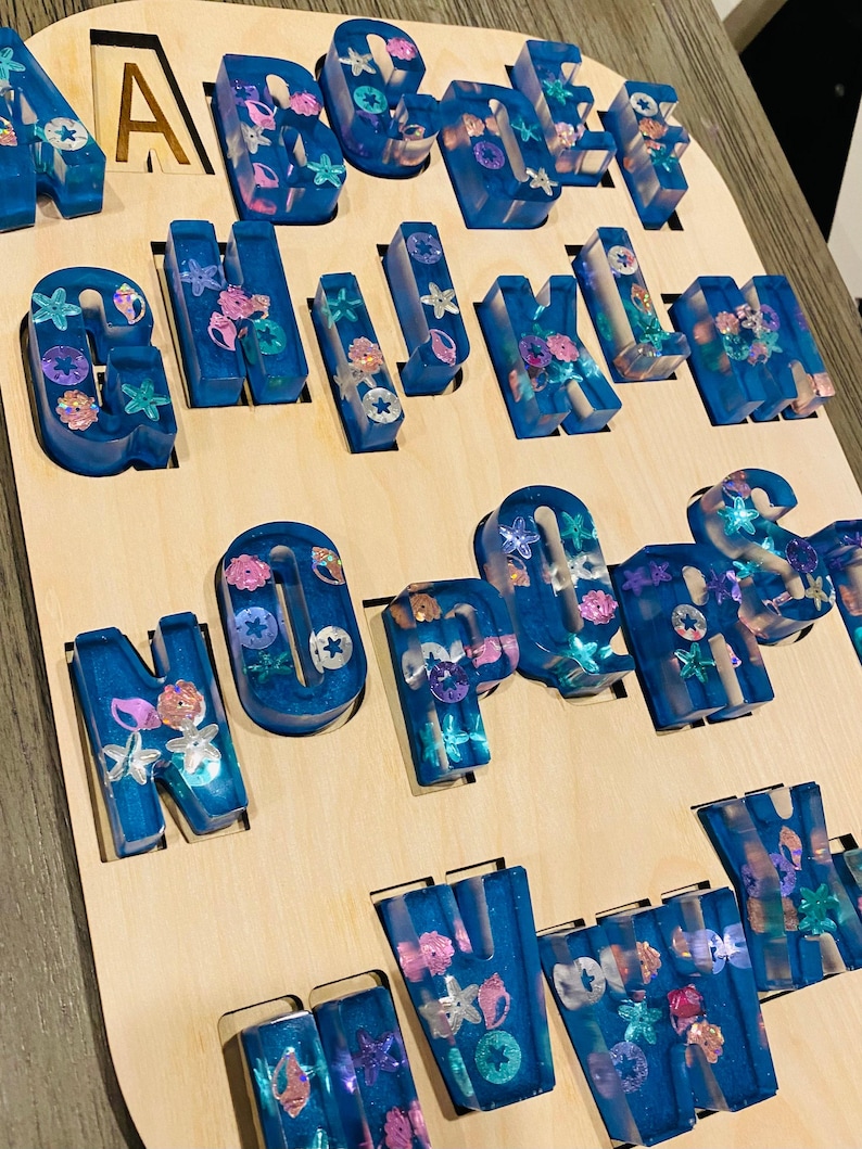 A wooden puzzle board with 26 chunky, translucent blue resin letters, each embedded with glitter and small seashell shapes, filling the letter-shaped cutouts.