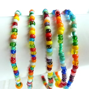 Könnte beinhalten: Eine Sammlung farbenfroher Perlenarmbänder. Jedes Armband besteht aus kleinen, runden Perlen in verschiedenen Farben, darunter Rot, Orange, Gelb, Grün, Blau und Lila. Die Armbänder sind gewickelt und scheinen flexibel zu sein.