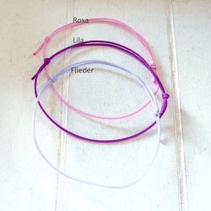 Könnte beinhalten: Drei farbige Armbänder in den Farben Rosa, Lila und Lavendel. Die Armbänder bestehen aus dünnem Kordel und sind mit einem Knoten gebunden. Die Wörter "Rosa", "Lila" und "Flieder" sind auf dem Bild zu sehen.