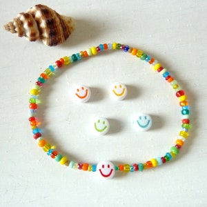 Puede incluir: Un brazalete de cuentas de colores con dijes de caritas sonrientes. El brazalete está hecho con cuentas pequeñas de varios colores y tiene cuatro dijes de caritas sonrientes blancas.