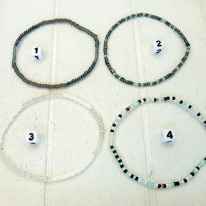 Könnte beinhalten: Vier Perlenarmbänder in verschiedenen Farben und Mustern. Die Armbänder sind quadratisch auf einer weißen Oberfläche angeordnet. Die Armbänder sind von 1 bis 4 nummeriert.
