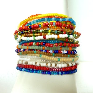 Op de afbeelding: Een stapel kleurrijke kralenarmbanden in tinten rood, geel, blauw, groen en wit. De armbanden zijn gemaakt van kleine, ronde kralen en zijn in een gelaagd patroon gerangschikt.