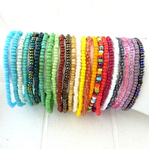 Könnte beinhalten: Eine Sammlung von 20 bunten Perlenarmbändern, jedes mit einem anderen Muster und einer anderen Farbkombination. Die Armbänder sind aus kleinen, runden Perlen in verschiedenen Blautönen, Grüntönen, Rottönen, Gelbtönen, Orangetönen, Weißtönen, Grautönen und Violetttönen gefertigt.