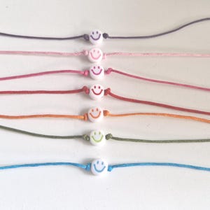 Könnte beinhalten: Sieben Armbänder in einer vertikalen Reihe, jedes mit einer einzelnen weißen runden Perle mit einem farbigen Smiley-Gesicht. Die Schnüre sind in den Farben Violett, Hellrosa, Dunkelrosa, Rot, Orange, Olivgrün und Hellblau gehalten.