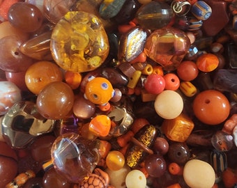 May include: A large assortment of colorful glass and ceramic beads in shades of orange, brown, and yellow. The beads are of various shapes and sizes, including round, square, and irregular.
