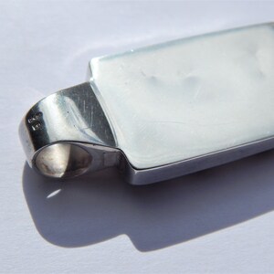 May include: Silver pendant with a rectangular shape and a smooth, polished surface. The pendant has a loop on the side for attaching a chain.