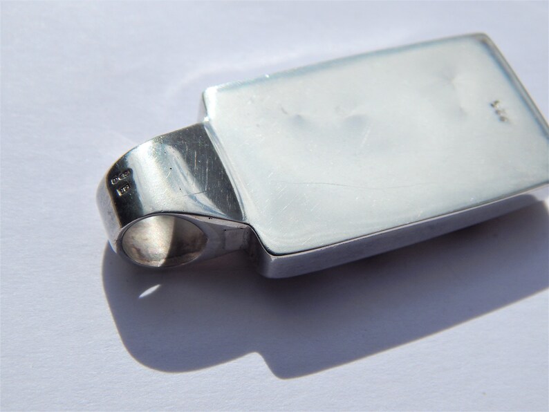 May include: Silver pendant with a rectangular shape and a smooth, polished surface. The pendant has a loop on the side for attaching a chain.