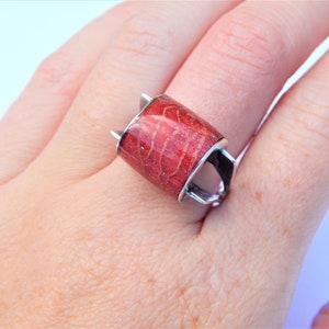 Puede incluir: Un anillo de plata con una piedra de coral rojo rectangular. El anillo tiene un dise&ntilde;o &uacute;nico con dos puntas en la parte superior y dos puntas en la parte inferior.