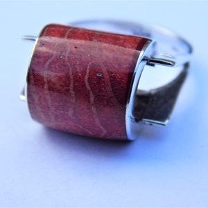 Puede incluir: Un anillo de plata con una piedra de coral rojo engastada en una banda de plata curvada y pulida. La piedra de coral presenta un patr&oacute;n natural y org&aacute;nico.