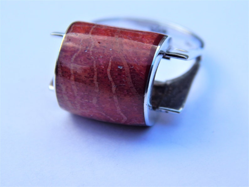 Puede incluir: Un anillo de plata con una piedra de coral rojo engastada en una banda de plata curvada y pulida. La piedra de coral presenta un patr&oacute;n natural y org&aacute;nico.