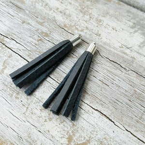 Puede incluir: Dos borlas de cuero negro con tapas de metal plateado. Las borlas miden aproximadamente 5 cm de largo y están colgando de una superficie de madera.