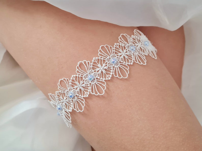 Op de afbeelding: Wit kanten kousenband met blauwe, iriserende kralen. Het kousenband is gemaakt van delicate kant en heeft een bloemenpatroon.