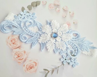 May include: A blue and white lace garter with a blue rhinestone embellishment. The garter is decorated with floral designs.