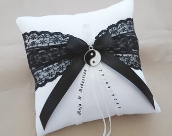 May include: White satin ring pillow with black lace trim and a black satin ribbon tied in a bow. A silver yin yang charm hangs from the bow. The pillow is personalized with the text "Anja & Sebastian" and the date "22.06.2019".