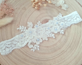May include: A white lace garter featuring intricate floral embroidery and small light blue pearl accents. The garter is constructed from a delicate, scalloped-edge lace band with a central applique of embroidered flowers and leaves.