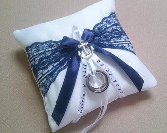 May include: White satin ring pillow with navy blue lace trim and a navy blue ribbon. The pillow has a silver anchor charm and two wedding bands. The ribbon is personalized with the text "Silvia & Jorg" and the date "05.08.2017".