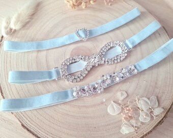 May include: Three light blue velvet garters with rhinestone embellishments. The garters feature a heart-shaped rhinestone, an infinity symbol, and a cluster of rhinestones.