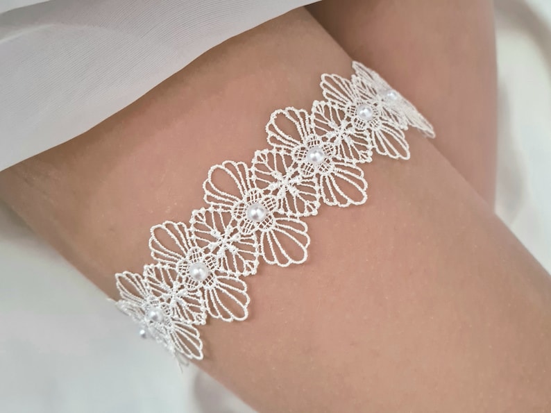 Op de afbeelding: Wit kanten kousenband met parelversiering. Het kousenband is gemaakt van delicate witte kant en heeft een rij parels.