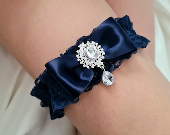 May include: A navy blue garter with a satin bow and a silver rhinestone embellishment. The embellishment features a large, clear rhinestone surrounded by a silver filigree design. A smaller, clear rhinestone dangles from the center of the embellishment.