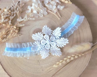 May include: A delicate, white lace garter with a light blue ribbon and a snowflake design. The garter features ruffled edges and is set against a rustic wooden background, with dried floral accents.