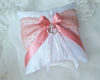 May include: White satin ring pillow with a pink lace trim and a pink satin bow. The pillow has a silver anchor charm and two white ribbons with the text "Lea & Alex" and "21.08.2021" in pink.