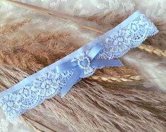 May include: A light blue lace garter with a delicate floral pattern. A matching blue satin bow is centered with a pearl heart. The garter is a wedding accessory, perfect for a bride.
