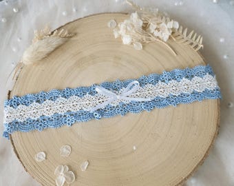 May include: A garter featuring a central band of white lace flanked by two rows of light blue floral crochet lace. The design is accented with a small, centered white satin ribbon bow. The lace construction displays intricate floral patterns.