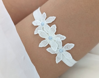 Könnte beinhalten: Weißes Spitzenstrumpfband mit drei blauen Blumenverzierungen. Das Strumpfband wird an einem Bein getragen.