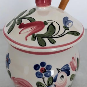 以下が含まれることがあります： ピンクとブルーの花柄が描かれた白い陶器製のハチミツポットと、木製のはちみつディッパー。ポットにはピンクのノブが付いた蓋が付いています。