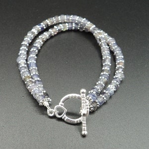 May include: A double-strand bracelet featuring small, faceted blue-gray beads. The bracelet is secured with a silver-toned toggle clasp, with a heart-shaped detail. The beads are strung on a flexible cord.
