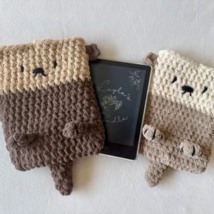 May include: Two rectangular crochet sleeves designed to look like otters, one in dark brown and tan, the other in light brown and cream. Each features small ears, embroidered faces, and paws. A black e-reader with the text "Cayla's Kindle" is centered.