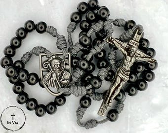 May include: A rosary featuring polished black beads strung on gray cord, a silver-toned crucifix with the letters I.N.R.I. at the top, and a shield-shaped Saint Benedict medal. The rosary is arranged in a circular pattern on a white surface.