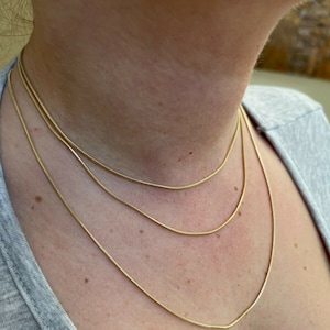 Puede incluir: Tres collares de cadena de oro superpuestos en el cuello de una persona. Las cadenas son finas y delicadas.