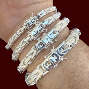 May include: Four silver bracelets with a detailed, ornate design are displayed. The bracelets are made of a shiny, metallic material and feature a repeating pattern of interlocking links. The bracelets are on a person's wrist.