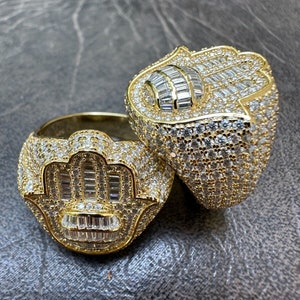 Puede incluir: Dos anillos de oro con un dise&ntilde;o de mano de hamsa, engastados con piedras de circonita c&uacute;bica transparentes. Los anillos est&aacute;n apilados uno encima del otro.