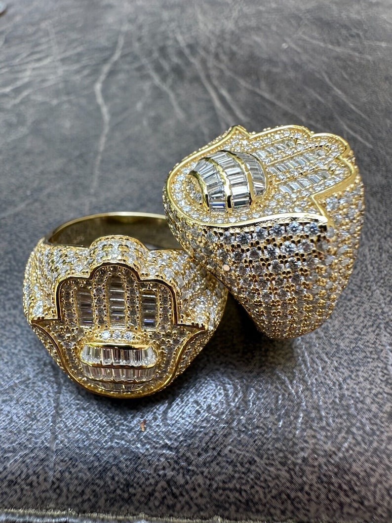 Puede incluir: Dos anillos de oro con un dise&ntilde;o de mano de hamsa, engastados con piedras de circonita c&uacute;bica transparentes. Los anillos est&aacute;n apilados uno encima del otro.