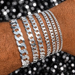 May include: A close-up of a wrist adorned with seven silver chain bracelets of varying widths. The bracelets have a curb link design and reflect light. The background is a sparkly, dark surface.