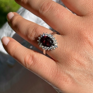 May include: A pear-shaped red gemstone ring with a halo of smaller clear gemstones. The ring is set in silver.
