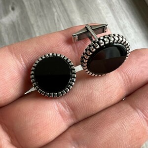 Op de afbeelding: Een paar zilveren manchetknopen met een zwarte, cirkelvormige middensteen. De manchetknopen hebben een decoratieve, getextureerde rand en een zilveren staafje voor bevestiging. De manchetknopen worden getoond op een lichtgrijze ondergrond.