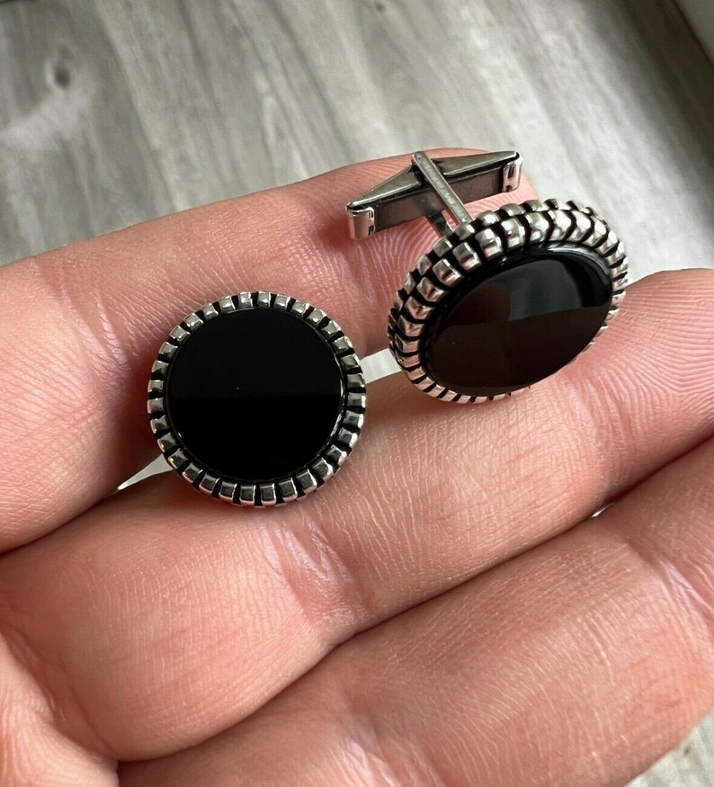 Op de afbeelding: Een paar zilveren manchetknopen met een zwarte, cirkelvormige middensteen. De manchetknopen hebben een decoratieve, getextureerde rand en een zilveren staafje voor bevestiging. De manchetknopen worden getoond op een lichtgrijze ondergrond.