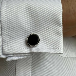 Op de afbeelding: Close-up van een wit overhemd met een textuurpatroon. De manchet van het overhemd heeft een zwarte, cirkelvormige manchetknop met een zilverkleurige, kralenrand. De manchetknop is gecentreerd op de manchet.