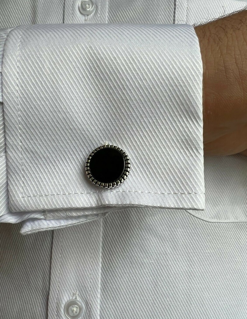 Op de afbeelding: Close-up van een wit overhemd met een textuurpatroon. De manchet van het overhemd heeft een zwarte, cirkelvormige manchetknop met een zilverkleurige, kralenrand. De manchetknop is gecentreerd op de manchet.