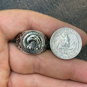 Puede incluir: Un anillo de plata con un dise&ntilde;o de cabeza de &aacute;guila. El anillo se muestra junto a una moneda de veinticinco centavos de d&oacute;lar estadounidense para comparar el tama&ntilde;o.