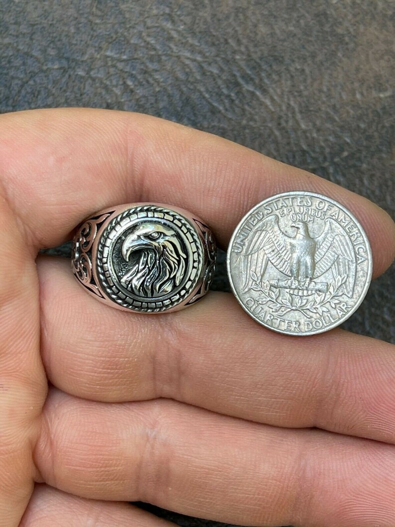 Puede incluir: Un anillo de plata con un dise&ntilde;o de cabeza de &aacute;guila. El anillo se muestra junto a una moneda de veinticinco centavos de d&oacute;lar estadounidense para comparar el tama&ntilde;o.