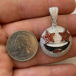 Puede incluir: Un colgante de plata con un dise&ntilde;o de cara sonriente. La cara sonriente tiene ojos en forma de estrella roja, una boca blanca y una nariz negra. El colgante est&aacute; cubierto de peque&ntilde;as piedras preciosas transparentes y rojas.